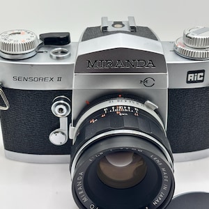 May include: A silver and black Miranda Sensorex II 35mm film camera with a black lens. The camera body features the text Miranda, Sensorex II, and AiC. The lens is marked with aperture and focus settings, and the text Lens Made in Japan.