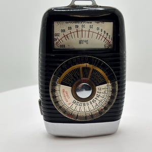 May include: A black DeJur Dual Professional light meter with a rectangular display showing numerical scales and a large circular dial featuring a silver center, black and white markings, and the text REFLECTED LIGHT printed in red on the face.