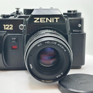 以下が含まれることがあります： 黒いZenit 122 35mmフィルムカメラで、目立つMC Helios 44M-4 2/58レンズが装着されています。カメラ本体には金色の文字でZenitのブランド名が表示され、前面にはモデル番号122が見えます。取り外された黒いレンズキャップが近くに置かれています。