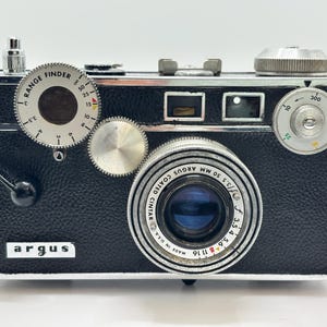 May include: A vintage Argus rangefinder camera with a black textured body and chrome accents. The lens is marked Argus Coated Cintar 50mm f/3.5 Made in U.S.A. It features a circular range finder dial, a shutter speed dial, and a viewfinder window.