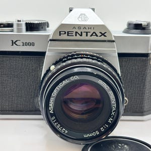 May include: An Asahi Pentax K1000 35mm film camera with a silver and black body. The camera features an SMC Pentax-M 1:2 50mm lens with the text Asahi Optical Co. printed around the front element. It includes metal strap lugs and a black lens cap.