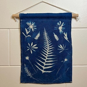 May include: A blue fabric wall hanging with white botanical prints of ferns and flowers. The fabric is hanging from a wooden dowel rod with white string.