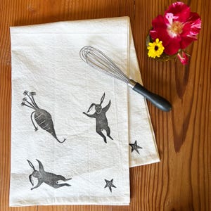 May include: White kitchen towel with black ink illustrations of a carrot, two rabbits, and two stars.