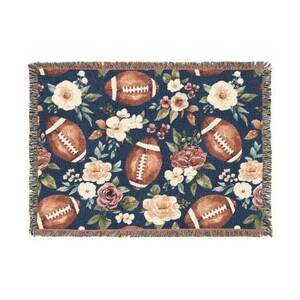 May include: A woven throw blanket with a navy blue background featuring a repeating pattern of brown American footballs and floral arrangements in cream, peach, and burgundy. The blanket has a fringed edge.