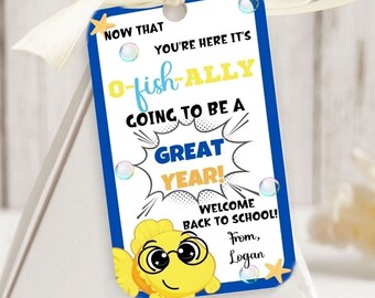 Op de afbeelding: Een blauw en geel terug naar school-label met een cartoonvis die een bril draagt. Het label zegt "Nu je hier bent, is het officieel een geweldig jaar! Welkom terug op school! Van Logan".