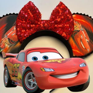 K&ouml;nnte beinhalten: Eine rote Lightning McQueen-Figur steht vor einem schwarzen Stirnband mit runden Ohren. Die Ohren zeigen eine Grafik von Lightning McQueen auf einem feurigen Hintergrund. Eine gro&szlig;e, paillettenbesetzte rote Schleife sitzt oben auf dem Stirnband.
