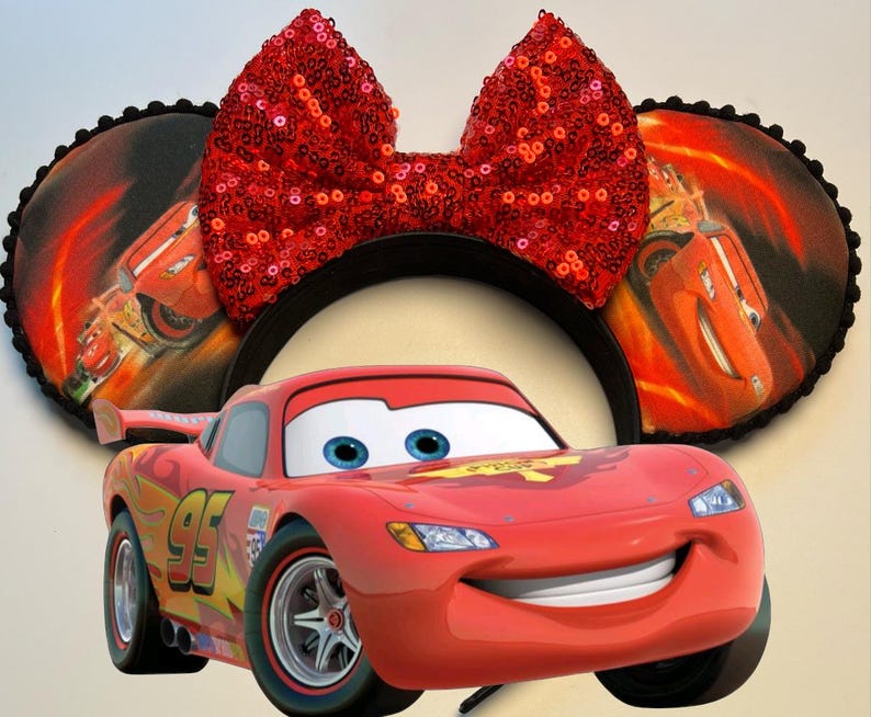 K&ouml;nnte beinhalten: Eine rote Lightning McQueen-Figur steht vor einem schwarzen Stirnband mit runden Ohren. Die Ohren zeigen eine Grafik von Lightning McQueen auf einem feurigen Hintergrund. Eine gro&szlig;e, paillettenbesetzte rote Schleife sitzt oben auf dem Stirnband.