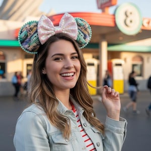 May include: A woman wearing a light blue denim jacket and a red and white striped shirt smiles at the camera. She is wearing a pair of mouse ears with a pink sequined bow and Cars-themed ear details. The background includes a cafe and people.
