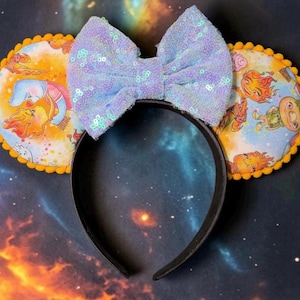 May include: A black headband featuring two circular ears with a yellow pom-pom trim. The ears display a colorful cartoon print of fire and water characters. A large, light blue sequined bow is centered between the ears on the headband.