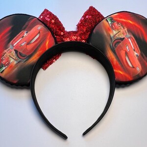 K&ouml;nnte beinhalten: Schwarzes Stirnband mit zwei runden Ohren mit einem roten Rennwagen-Design auf einem feurigen Hintergrund. Eine rote Pailletten-Schleife befindet sich zwischen den Ohren. Die Ohren sind mit schwarzem Paspel verziert.