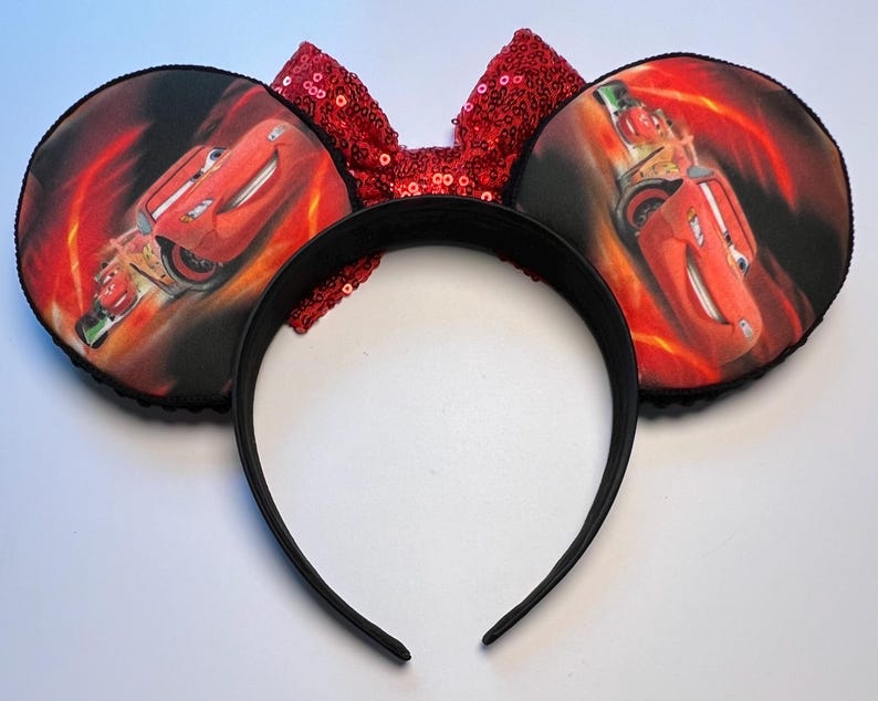 K&ouml;nnte beinhalten: Schwarzes Stirnband mit zwei runden Ohren mit einem roten Rennwagen-Design auf einem feurigen Hintergrund. Eine rote Pailletten-Schleife befindet sich zwischen den Ohren. Die Ohren sind mit schwarzem Paspel verziert.