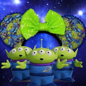 May include: A Disney-themed headband featuring three green alien figures with three eyes each, wearing blue and purple outfits. The headband has blue ears with an alien pattern and a bright green sequined bow.