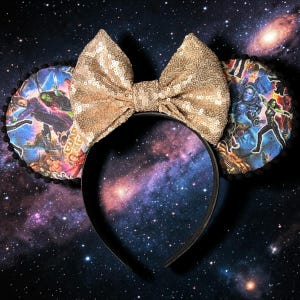 May include: A headband featuring two circular ears decorated with a colorful Guardians of the Galaxy character print and black pom-pom trim. A large, central bow covered in shimmering gold sequins sits between the ears on a black plastic band.