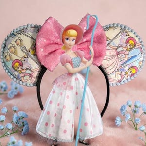 May include: A Bo Peep-themed headband with a large pink sequined bow and two circular ear designs. The ears feature illustrations of Bo Peep and sheep. A Bo Peep figurine is in the center, holding a blue shepherd's crook. The headband is black.