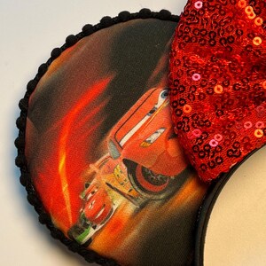 K&ouml;nnte beinhalten: Mausohren mit einer roten Pailletten-Schleife und einem kreisf&ouml;rmigen Ohr mit einer Grafik eines roten Rennwagens. Das Ohr ist mit schwarzen Pompons eingefasst und an einem schwarzen Stirnband befestigt. Das Auto ist vor einem feurigen orange-roten Hintergrund platziert.