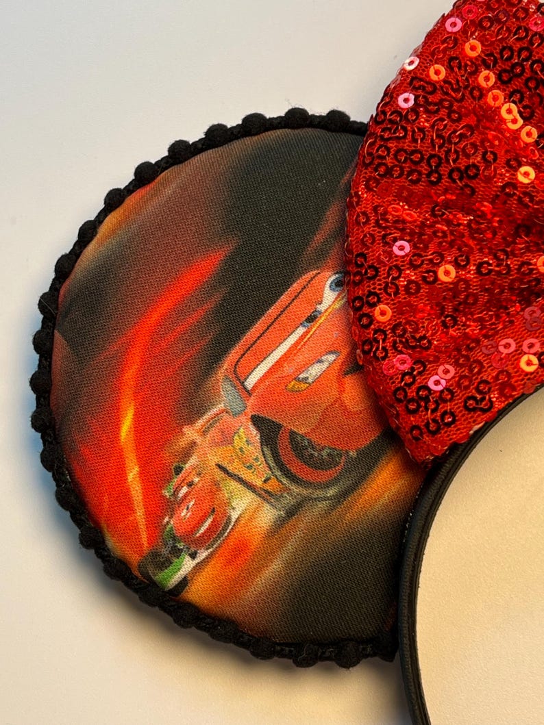 K&ouml;nnte beinhalten: Mausohren mit einer roten Pailletten-Schleife und einem kreisf&ouml;rmigen Ohr mit einer Grafik eines roten Rennwagens. Das Ohr ist mit schwarzen Pompons eingefasst und an einem schwarzen Stirnband befestigt. Das Auto ist vor einem feurigen orange-roten Hintergrund platziert.