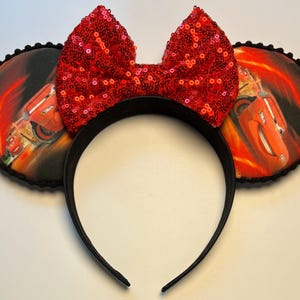 K&ouml;nnte beinhalten: Schwarzes Stirnband mit zwei runden Ohren mit einem Cars-Filmmotiv. Die Ohren haben eine schwarze Umrandung und ein Bild von Lightning McQueen. Eine gro&szlig;e rote Pailletten-Schleife ist mittig auf dem Stirnband angebracht und sorgt f&uuml;r einen Hauch von Glanz.