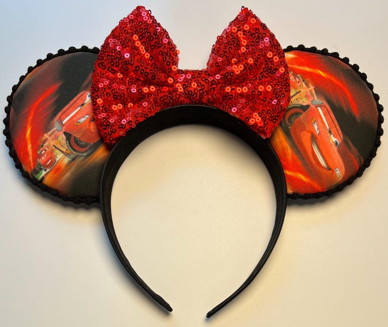 K&ouml;nnte beinhalten: Schwarzes Stirnband mit zwei runden Ohren mit einem Cars-Filmmotiv. Die Ohren haben eine schwarze Umrandung und ein Bild von Lightning McQueen. Eine gro&szlig;e rote Pailletten-Schleife ist mittig auf dem Stirnband angebracht und sorgt f&uuml;r einen Hauch von Glanz.