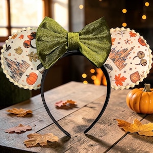 May include: A headband featuring two circular ears with a cream-colored fabric printed with autumn-themed illustrations including pumpkins, leaves, and castles. A large, shimmering olive green velvet bow sits between the ears, which are trimmed with small white pom-poms.
