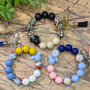 May include: Three colorful beaded keychains with a Mickey Mouse head charm. Each keychain has a different pattern and color scheme: black and white animal print, blue and white floral, and pink and blue floral. The keychains are made of silicone beads and have a silver key ring.