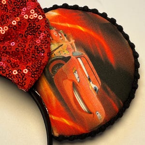 K&ouml;nnte beinhalten: Schwarzes Stirnband mit zwei runden Ohren mit einem Cars-Film-Motiv. Die Ohren sind mit schwarzen Pompons verziert. Eine gro&szlig;e rote Pailletten-Schleife ist am Stirnband befestigt. Die Cars-Grafik zeigt Lightning McQueen.