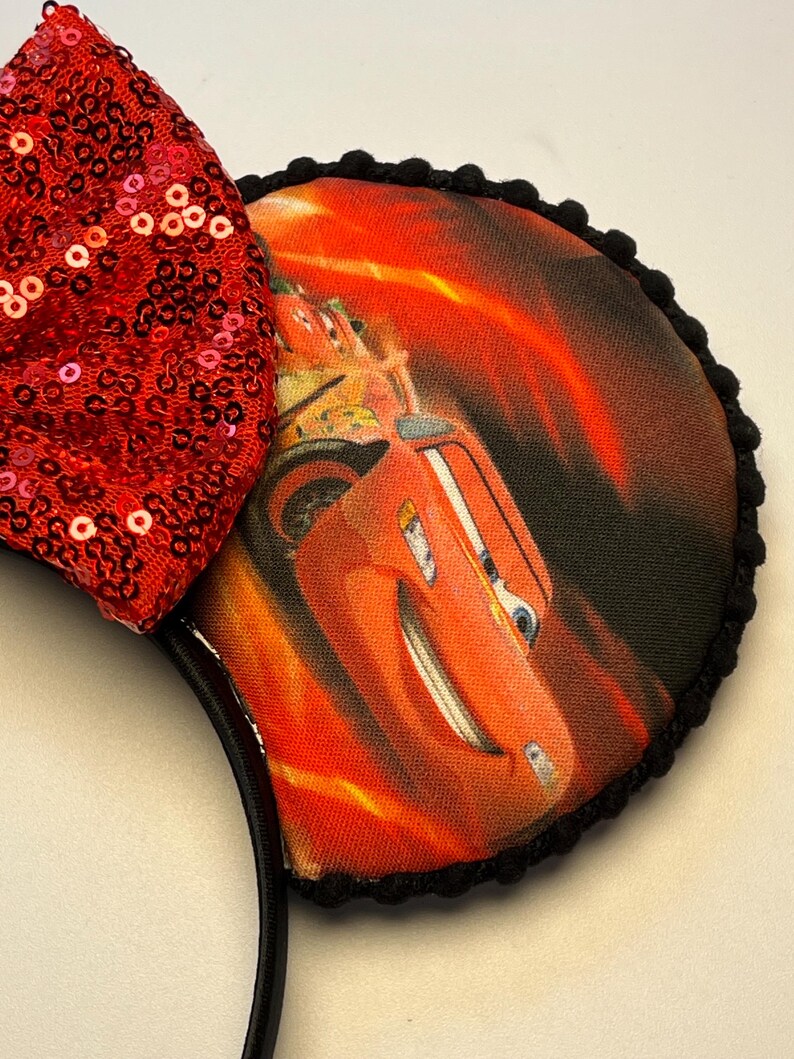 K&ouml;nnte beinhalten: Schwarzes Stirnband mit zwei runden Ohren mit einem Cars-Film-Motiv. Die Ohren sind mit schwarzen Pompons verziert. Eine gro&szlig;e rote Pailletten-Schleife ist am Stirnband befestigt. Die Cars-Grafik zeigt Lightning McQueen.