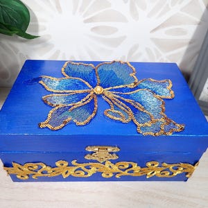 Op de afbeelding: Een rechthoekige houten doos geschilderd in een levendige koningsblauwe kleur. Het deksel is voorzien van een grote, gedetailleerde blauwe bloemapplicatie omlijnd met glinsterende gouden lovertjes en een centrale parel. Een gouden sluiting en gouden rand versieren de voorzijde.