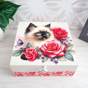 Op de afbeelding: Een vierkante witte houten doos met een kleurrijke illustratie op het deksel van een pluizige kat met blauwe ogen, omringd door levendige rode rozen, kleine paarse bloemen en een blauwe vlinder. Het voorpaneel toont een rood bloemmotief.