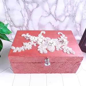 May include: A rectangular rose gold jewellery box with a textured leaf pattern on the sides and a clear crystal knob. The lid is decorated with a white lace floral appliqué featuring small white pearls centred on the petals and leaves.
