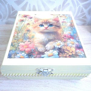 May include: A rectangular wooden box with a light cream finish and a decorative silver-toned metal latch. The lid features a printed illustration of a fluffy ginger kitten with bright blue eyes sitting among a field of colourful wildflowers.