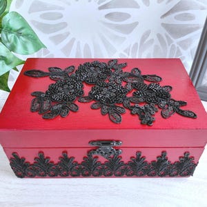 May include: A rectangular red wooden box featuring a black sequined floral applique on the lid and a black lace trim along the front edge. The box includes a metal latch closure on the front and has a smooth, painted finish on all visible sides.
