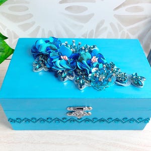 Op de afbeelding: Een rechthoekige turquoise houten doos met een zilverkleurige metalen sluiting aan de voorzijde. Het deksel is versierd met een cluster van blauwe stoffen bloemen, heldere kristallen en lovertjes. Een rij blauwe strass-steentjes siert de rand.