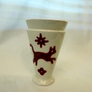 May include: Cream-colored ceramic vase with a flared top and a footed base. The vase is decorated with a dark red cat, star, and flower design. The vase has a smooth, glossy finish and is suitable for displaying flowers or as a decorative item.