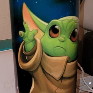 May include: A green and brown character with large ears and big eyes is printed on a black and white tumbler. The background is a dark blue with green and white dots.