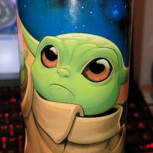 May include: A green and brown character with large eyes and a brown collar is printed on a black and blue tumbler with green glitter. The tumbler is sitting on a white coaster.