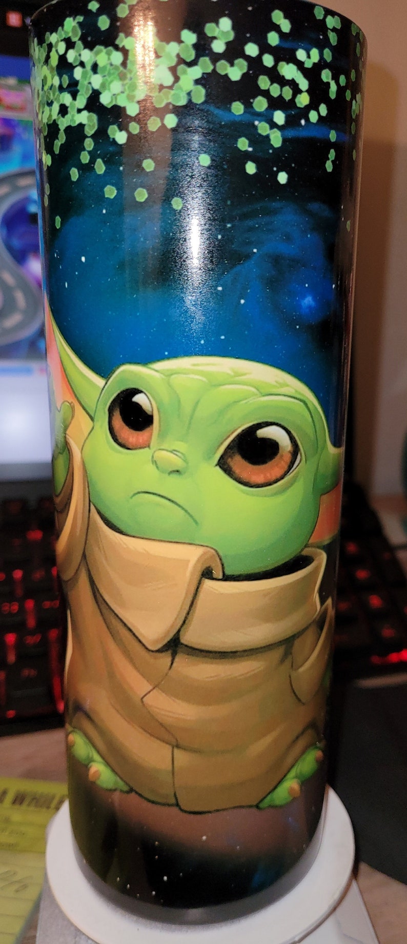 May include: A green and brown character with large eyes and a brown collar is printed on a black and blue tumbler with green glitter. The tumbler is sitting on a white coaster.