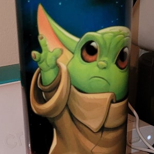 May include: A green and brown character with large ears and big eyes is printed on a black and white tumbler. The background is a dark blue with green and white dots.