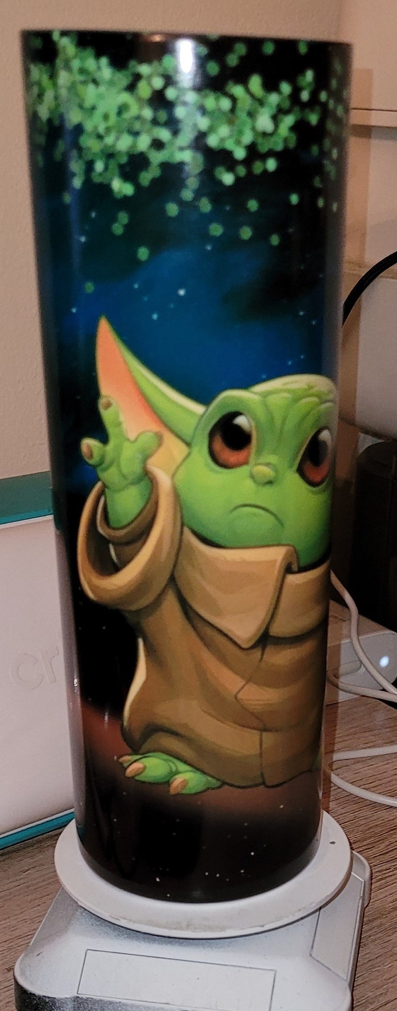 May include: A green and brown character with large ears and big eyes is printed on a black and white tumbler. The background is a dark blue with green and white dots.