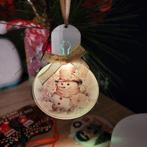 Könnte beinhalten: Ein runder weißer Weihnachtsbaumschmuck mit einem Schneemann-Motiv und einem goldenen Band. Der Schmuck hat ein Licht im Inneren, das den Schneemann beleuchtet.