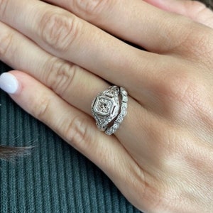 May include: A close-up of a woman's hand wearing a diamond engagement ring and a wedding band. The ring is set in a platinum band and features a large, square-cut diamond.