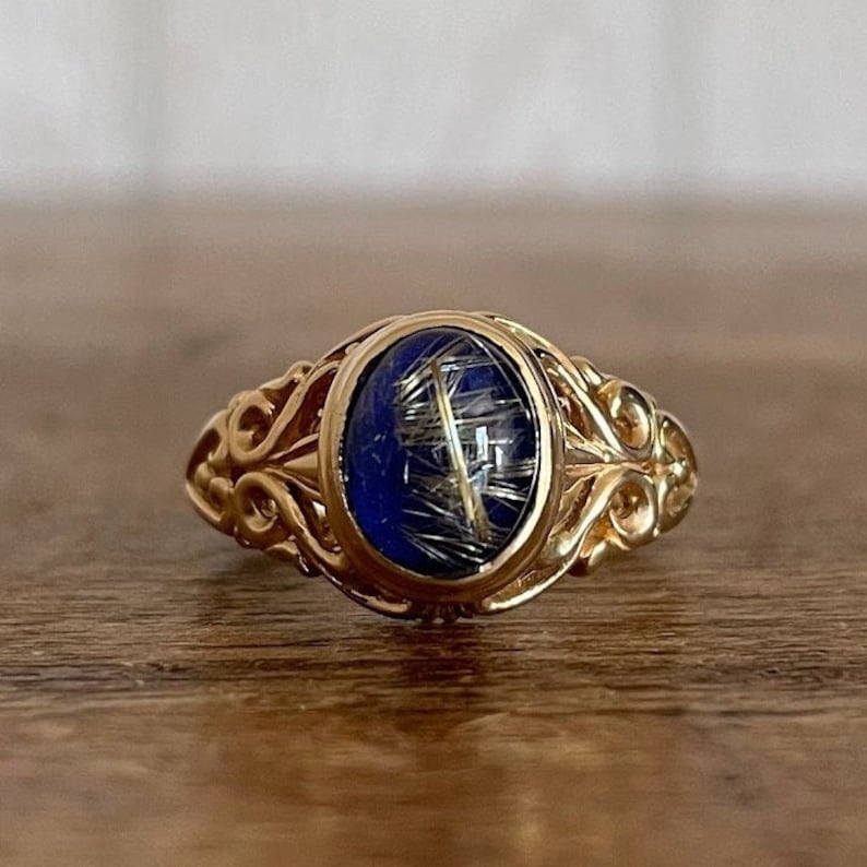 May include: A gold ring with an oval-shaped blue stone set in a filigree design. The stone has a gold-colored inclusion.