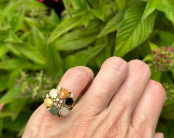 Könnte beinhalten: Ein goldener Ring mit einem Blumendesign mit sieben Edelsteinen. Die Edelsteine sind grün, weiß, schwarz, orange und gelb.