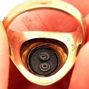 K&ouml;nnte beinhalten: Nahaufnahme des Inneren eines Goldrings mit zwei kleinen schwarzen Punkten und der Inschrift "M" an der Seite.