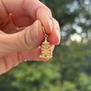 May include: A gold-colored charm in the shape of a pagoda, a traditional East Asian temple. The charm is being held by a hand against a blurred green background.