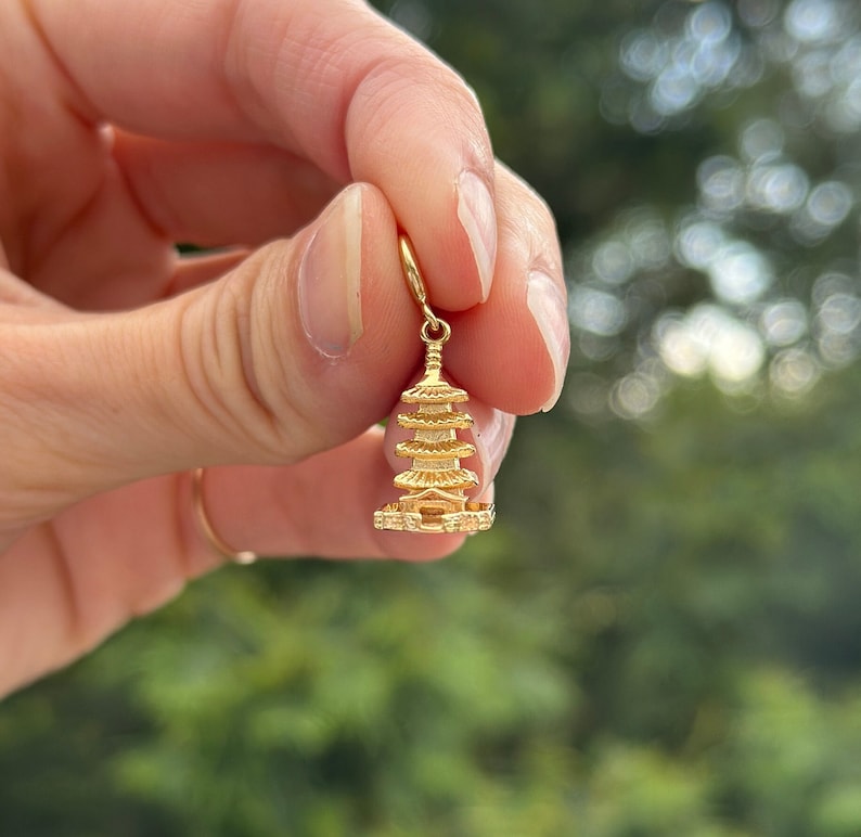 May include: A gold-colored charm in the shape of a pagoda, a traditional East Asian temple. The charm is being held by a hand against a blurred green background.