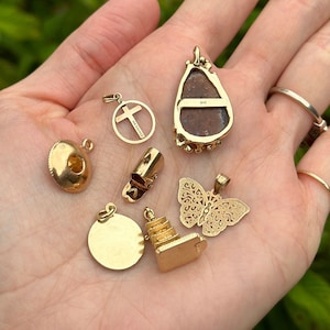 May include: A collection of gold-toned charms, including a cross, a butterfly, a pagoda, a round charm, and a teardrop-shaped charm with a brown stone. The charms are on a white background.