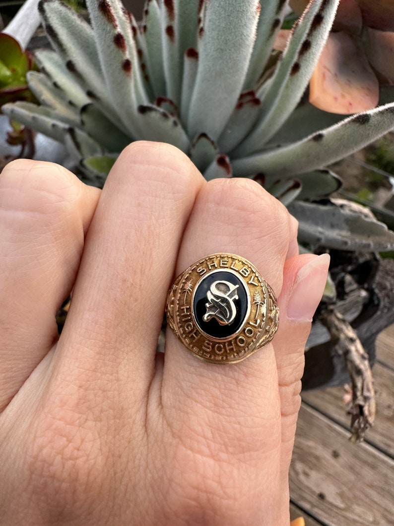 K&ouml;nnte beinhalten: Ein goldener Klassenring mit schwarzer Emaille-Einlage, die den Buchstaben "S" und die Worte "Shelby High School" zeigt.