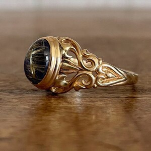 May include: A gold ring with an ornate design and a large, round, dark gemstone set in the center.