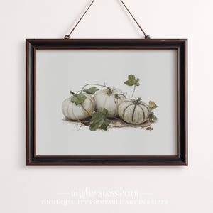 Peut inclure: Une impression encadrée présentant une illustration à l'aquarelle de trois citrouilles blanches avec des tiges et des feuilles vertes. Les citrouilles sont disposées sur un fond clair, avec un cadre marron foncé et une corde de suspension.
