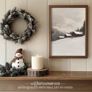 Peut inclure: Une impression encadrée d'un paysage d'hiver avec une scène de montagne enneigée et de petites maisons en bois. Une figurine de bonhomme de neige est posée sur une étagère en bois avec une bougie blanche et une couronne de verdure givrée.